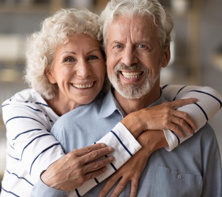 Head shot portrait of affectionate loving middle aged hoary beautiful woman cuddling from back smiling old husband. Happy loving mature married family couple looking at camera, posing for photo.